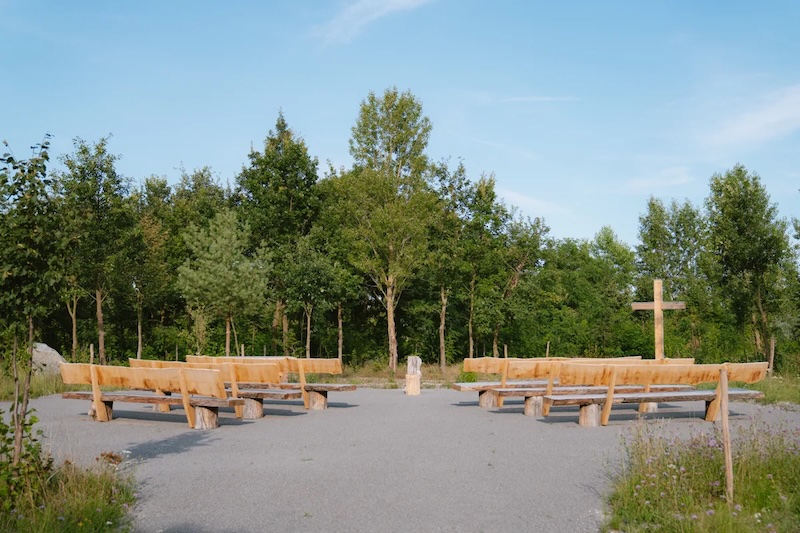 Andachtsplatz im Bestattungswald am Schladitzer See, die Holzbänke sind in einem Halbkreis um einen Urnenstele angeordnet, ein großes Holzkreuz steht seitlich des Andachtsplatzes